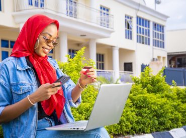 african-female-sitting-outside-with-her-laptop-phone-making-payment-online