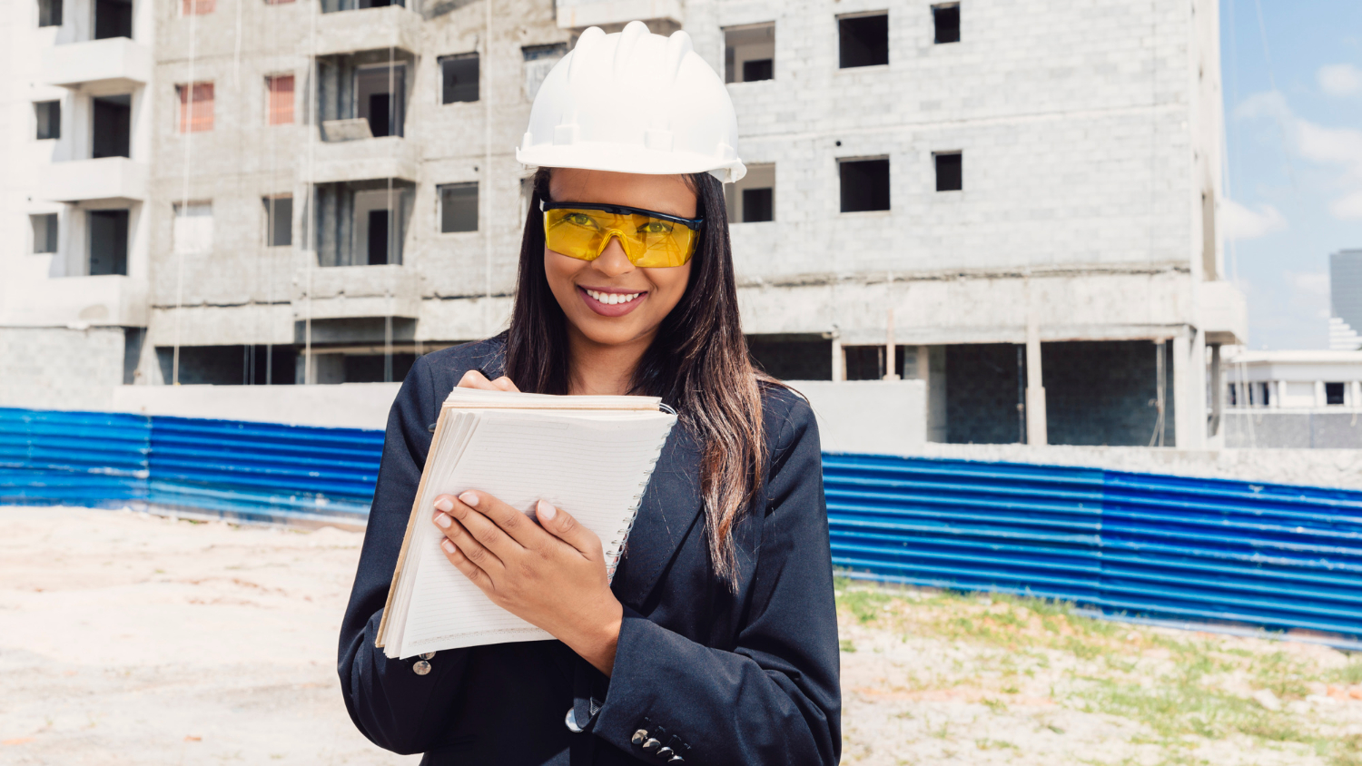 happy-african-american-lady-safety-helmet-writing-notebook-near-building-construction